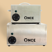 Two stacked rectangular brushed-metal heating units on small black feet; each unit has a black circular dial with white tick marks and small unreadable numbers on the lower left, a small round green indicator light above the dial, and the top unit has a white handle on top with white side handles; beige background. Text reads: ONCE NATURAL WAX; ONCE NATURAL WAX.